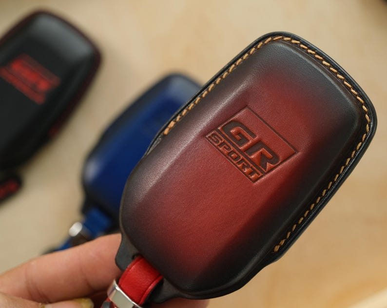 May include: A dark red and black gradient leather key fob cover featuring the embossed text GR SPORT. The cover has visible tan stitching along the edges and is attached to a red leather strap with a silver metal connector at the base.