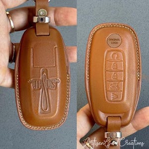 May include: A brown leather key fob case with a silver-tone metal ring. The front features a cross design with the word "JESUS". The back has buttons and the text "ORIGINAL LOGO".