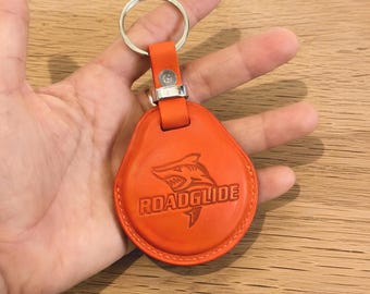 May include: A bright orange leather keychain featuring a circular shape with a metal split ring. The leather surface is embossed with a graphic of a shark and the text ROADGLIDE. The keychain has a stitched border and a silver metal connector.