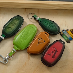 Puede incluir: Una colección de fundas de cuero para llaves de coche en verde oscuro, verde lima, tostado y rojo. La funda tostada presenta un diseño de corazón y cruz con el texto Jesus, mientras que la roja muestra una letra A grande. Etiquetas de cuero pequeñas están marcadas con Maxine, Alize, J.L.S, Billie y Hans-Peter.