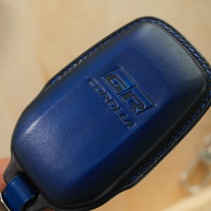 May include: A dark blue leather key fob cover featuring the embossed GR COROLLA logo on the front surface. The cover is constructed with matching blue stitching along the edges and includes a metal attachment loop at the base for a keychain.