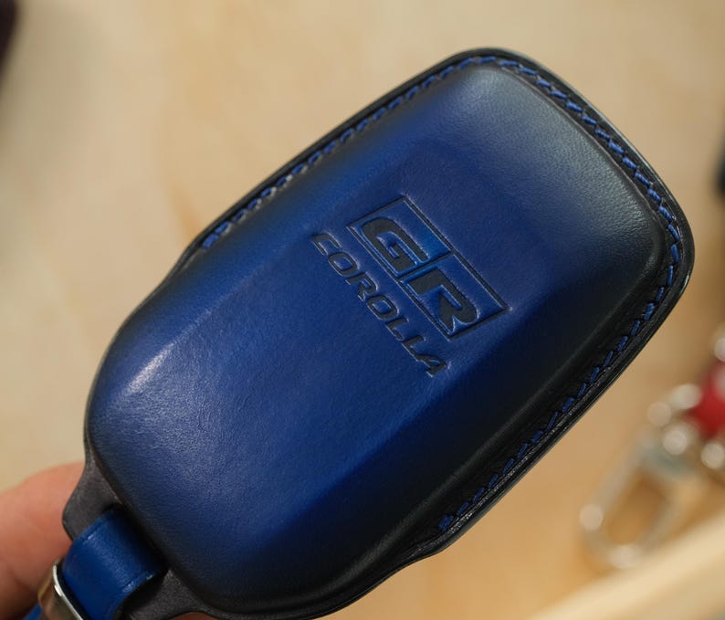 May include: A dark blue leather key fob cover featuring the embossed GR COROLLA logo on the front surface. The cover is constructed with matching blue stitching along the edges and includes a metal attachment loop at the base for a keychain.