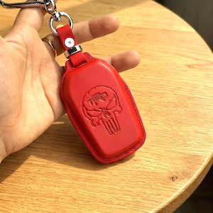 May include: A red leather key fob case with a silver keyring. The case features an embossed skull design with the letters "TRD". The key fob is held by a hand, and the background is a wooden surface.