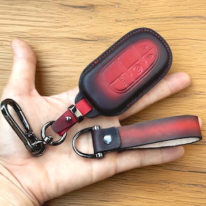 May include: A red and black leather key fob and matching wrist strap. The key fob has a red interior and a black exterior with red stitching. The wrist strap is red and black with a silver-tone metal D-ring and a black metal clasp.