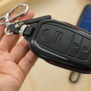May include: A black leather car key fob cover featuring red stitching along the edges. The cover has molded buttons for lock, unlock, and hold functions. It is attached to a silver-toned metal carabiner clip by a short black leather strap.
