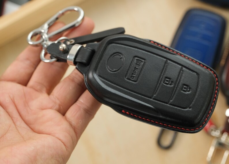 May include: A black leather car key fob cover featuring red stitching along the edges. The cover has molded buttons for lock, unlock, and hold functions. It is attached to a silver-toned metal carabiner clip by a short black leather strap.