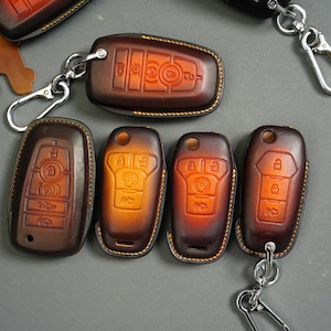 May include: A collection of brown leather key fob covers with silver keychains. The covers have embossed designs and are in various shades of brown with yellow stitching. The key fobs are for a car.