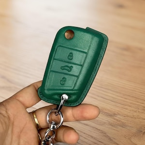 May include: A green leather car key fob cover with a silver key ring. The cover features three embossed buttons and a hole for the key. The key fob is green and has a stitched border.