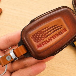 Peut inclure: Un étui à clé en cuir marron avec un motif de drapeau américain gaufré et le texte 4RUNNER. L'étui comprend un anneau métallique et une lanière en cuir fixée par un rivet argenté. Deux petites étiquettes rectangulaires en cuir sont visibles.