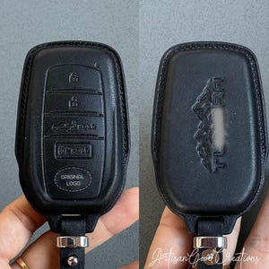 May include: Black leather car key fob cover with embossed buttons and the words "ORIGINAL LOGO". The back of the cover has an embossed design. The key fob cover is attached to a black leather strap with a silver-colored metal clasp.