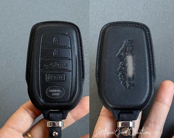 May include: Black leather car key fob cover with embossed buttons and the words "ORIGINAL LOGO". The back of the cover has an embossed design. The key fob cover is attached to a black leather strap with a silver-colored metal clasp.