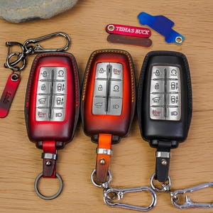 May include: Three leather car key fobs in red, orange, and black, each with silver key rings and keychains. The fobs have silver buttons and a leather strap. A red keychain with the name "Ludo" is attached to one fob.