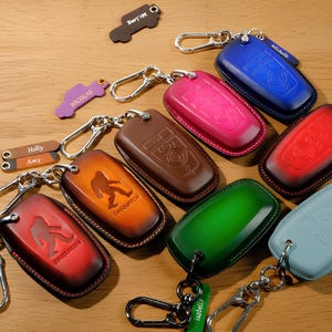 May include: A collection of leather keychains in various colors, including red, blue, green, and brown. Some keychains feature embossed designs, such as a Sasquatch figure. Each keychain has a metal clasp and a small tag with a name.