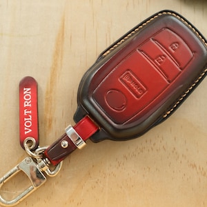 May include: A red and dark brown leather car key fob cover with visible stitching. The cover features molded button icons and is attached to a red leather strap with a silver metal clasp and a red tag with the white text VOLT RON printed on it.