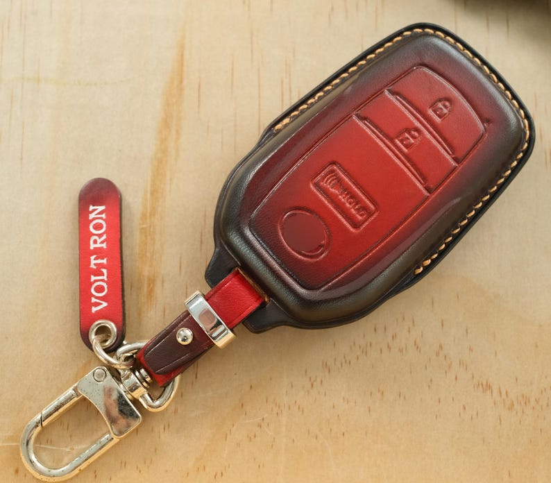 May include: A red and dark brown leather car key fob cover with visible stitching. The cover features molded button icons and is attached to a red leather strap with a silver metal clasp and a red tag with the white text VOLT RON printed on it.