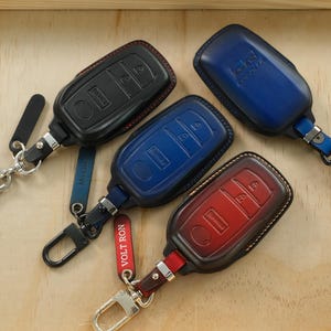 May include: Four leather car key fob covers in black, blue, and red, each featuring a metal carabiner clip and a small leather tag. The covers have molded button outlines and visible stitching. One red tag is printed with the text VOLT RON.