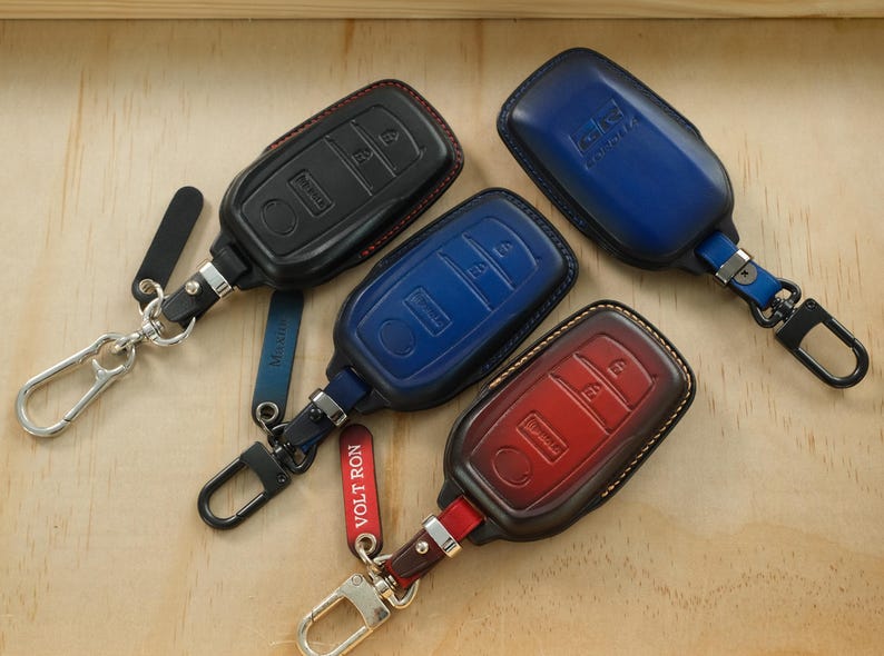 May include: Four leather car key fob covers in black, blue, and red, each featuring a metal carabiner clip and a small leather tag. The covers have molded button outlines and visible stitching. One red tag is printed with the text VOLT RON.