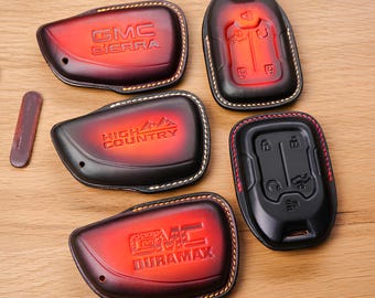 May include: A collection of leather key fob covers in red and black, featuring embossed GMC logos and vehicle model names like Sierra, High Country, and Duramax. The covers have contrasting stitching and a sleek design.