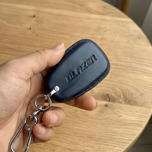 May include: A navy blue leather key fob with the word "BLAZER" embossed on the front. The key fob is attached to a silver metal keyring and a large carabiner clip.