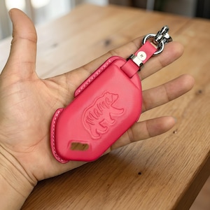 May include: A bright pink leather key fob with a silver-tone clasp. The fob features an embossed bear design with the word "Mama" and a small rectangular cutout. The key fob is held in a hand.