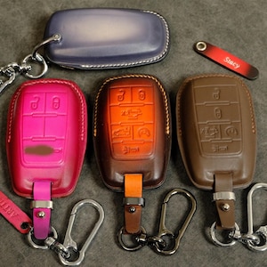 May include: A collection of leather key fob covers in various colors, including pink, brown, and blue. Each cover features a different design and includes a metal clasp for attachment. One tag reads "Stacy" and another reads "ARTISAN".