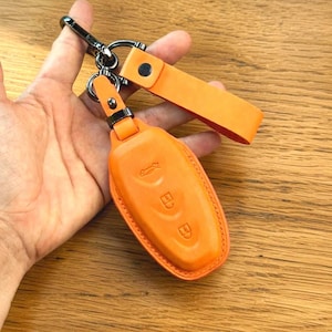 Könnte beinhalten: Orangenes Leder-Schlüsselanhänger-Etui mit passendem Riemen und silberfarbenem Metallverschluss. Das Etui hat geprägte Schlüsselsymbole und einen Knopfverschluss. Der Schlüsselanhänger dient zum Schutz von Autoschlüsseln.