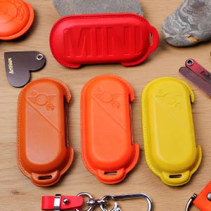 May include: A collection of four oval-shaped key fob covers in red, orange, and yellow. The top red cover features the embossed text MINI. The three lower covers display an embossed wing logo. Each cover includes a small hole for a keychain attachment.
