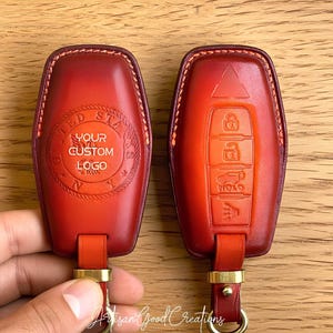 May include: Two red leather key fobs with gold hardware. One fob has the text "YOUR CUSTOM LOGO" inside a circular design. The other fob has embossed key symbols. The fobs are on a wooden surface.