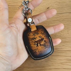 May include: A brown and black leather key fob with a dinosaur design. The fob has a metal keyring and a leather strap with a silver button. The edges are stitched with white thread.