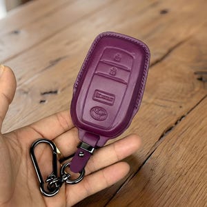 Può includere: Una custodia per portachiavi in pelle color prugna con moschettone e portachiavi in metallo nero. La custodia presenta icone in rilievo e un logo Toyota. Il portachiavi è tenuto in mano su uno sfondo di legno.