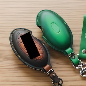 以下が含まれることがあります： 緑と茶色の革製キーフォブカバーのクローズアップ。緑色のカバーにはストラップとシルバーの金具が付いています。茶色のカバーには革のストラップと金属製の留め金が付いています。どちらも楕円形で、手作りのようです。
