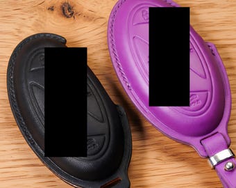 May include: Two oval-shaped key fob covers, one in black and one in purple, featuring molded button icons and stitched edges. The purple cover includes a silver-toned metal attachment loop and a small rivet, while both covers have a smooth finish.