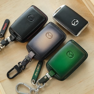 Puede incluir: Tres fundas de cuero para llaves de vehículos Mazda en negro, marrón oscuro y verde, cada una con el logotipo de Mazda en relieve. Las fundas incluyen anillas metálicas y etiquetas de cuero con el texto Maryin y JGo DB12, más una llave sin funda.