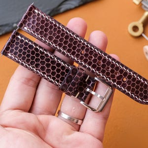 以下が含まれることがあります： 表面にテクスチャ加工されたハニカム模様が施されたダークブラウンの時計ベルト。ベルトの縁には白いコントラストステッチが入り、片端には固定用の磨かれたシルバーカラーの金属製バックルが取り付けられています。