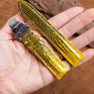 Könnte beinhalten: Zweiteiliges Uhrenarmband in leuchtendem Gelb mit Reptilienmuster. Das Armband wird in einer Hand gehalten. Es hat eine silberfarbene Schnalle. Der Hintergrund ist braun. Das Armband ist neu.