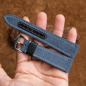 May include: Two-piece watch strap set in denim blue fabric with white stitching. The straps feature black leather accents and silver-tone buckles. The watch straps are held in a hand against a brown background.