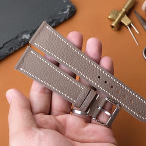May include: A taupe-colored leather watch strap featuring a textured grain surface and contrasting white stitching. The strap includes a silver-toned metal buckle and multiple adjustment holes, designed for secure attachment to a timepiece.