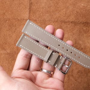 Puede incluir: Una correa de reloj de color topo claro con costuras blancas y una hebilla plateada. La correa está hecha de un material texturizado y tiene varios agujeros para ajustar. La correa del reloj se muestra sobre un fondo marrón.