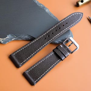 Può includere: Un cinturino per orologio in pelle marrone scuro con superficie testurizzata e cuciture bianche a contrasto. Il cinturino presenta una fibbia in metallo color argento e due passanti in pelle abbinati per il fissaggio alla cassa.