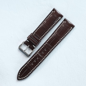 Könnte beinhalten: Ein dunkelbraunes Uhrenarmband mit geprägtem Alligator-Muster und kontrastierenden weißen Nähten an den Rändern. Das Armband verfügt über eine polierte silberfarbene Metallschnalle und zwei verschiebbare Schlaufen zur sicheren Befestigung.