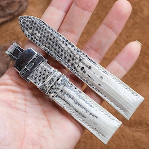 Puede incluir: Un par de correas de reloj con un patrón de piel de reptil en tonos grises y blancos. Las correas tienen un cierre de acero inoxidable y se muestran sobre un fondo marrón.