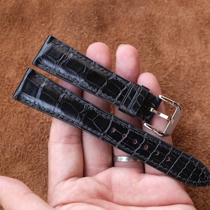 May include: A pair of black watch straps with a crocodile skin pattern. The straps are held in a hand against a brown background. The straps have silver-colored buckles.