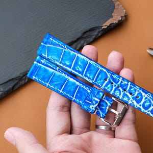 May include: A bright blue watch strap featuring a textured crocodile skin pattern with white stitching along the edges. The strap includes a polished silver-tone metal buckle and multiple adjustment holes for securing the watch to a wrist.