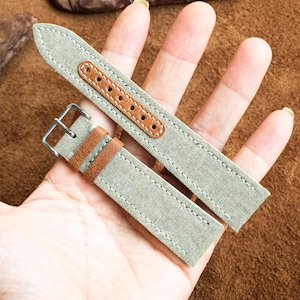 Op de afbeelding: Een paar horlogebandjes met een lichtgroen canvas buitenkant en bruine leren accenten. De bandjes hebben een gestikt ontwerp en een zilveren gesp. De horlogebandjes worden getoond tegen een bruine achtergrond.