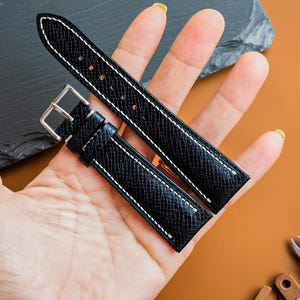 Puede incluir: Una correa de reloj negra con superficie texturizada y costuras blancas en contraste. La correa cuenta con una hebilla de metal plateado y varios orificios de ajuste, presentando un diseño refinado para diversos relojes.
