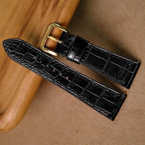 May include: A black watch strap with a glossy crocodile-embossed texture and a polished gold-tone metal buckle. The two-piece strap features a pointed tip on one end and a rectangular buckle with a metal pin on the other, showcasing a classic design.