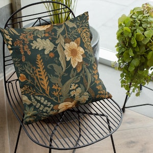 May include: Square decorative pillow with a botanical floral pattern in shades of teal, beige, and rust. The pillow is placed on a black metal chair. The design features flowers, leaves, and foliage.