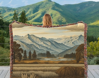 Può includere: Una coperta tessuta con un dipinto di paesaggio di montagne, alberi e un fiume. La coperta ha un bordo con frange con colori rosso, marrone e beige. L'opera d'arte raffigura una scena all'aperto serena con una tavolozza di colori tenui.