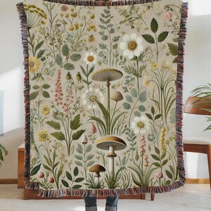May include: A beige woven throw blanket featuring a botanical design with mushrooms, flowers, and foliage in shades of green, yellow, pink, and white. The blanket has a fringed edge with red, blue, and brown threads. The blanket is approximately 60 inches wide and 80 inches long.