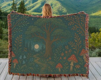 May include: A teal woven throw blanket featuring a forest scene with a large tree, mushrooms, and a full moon. The blanket has a fringed edge with shades of red, orange, and brown. The design includes a path leading to the moon.